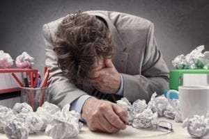 Exhausted man holding his head. He is surrounded by binders of documents and wadded up papers.