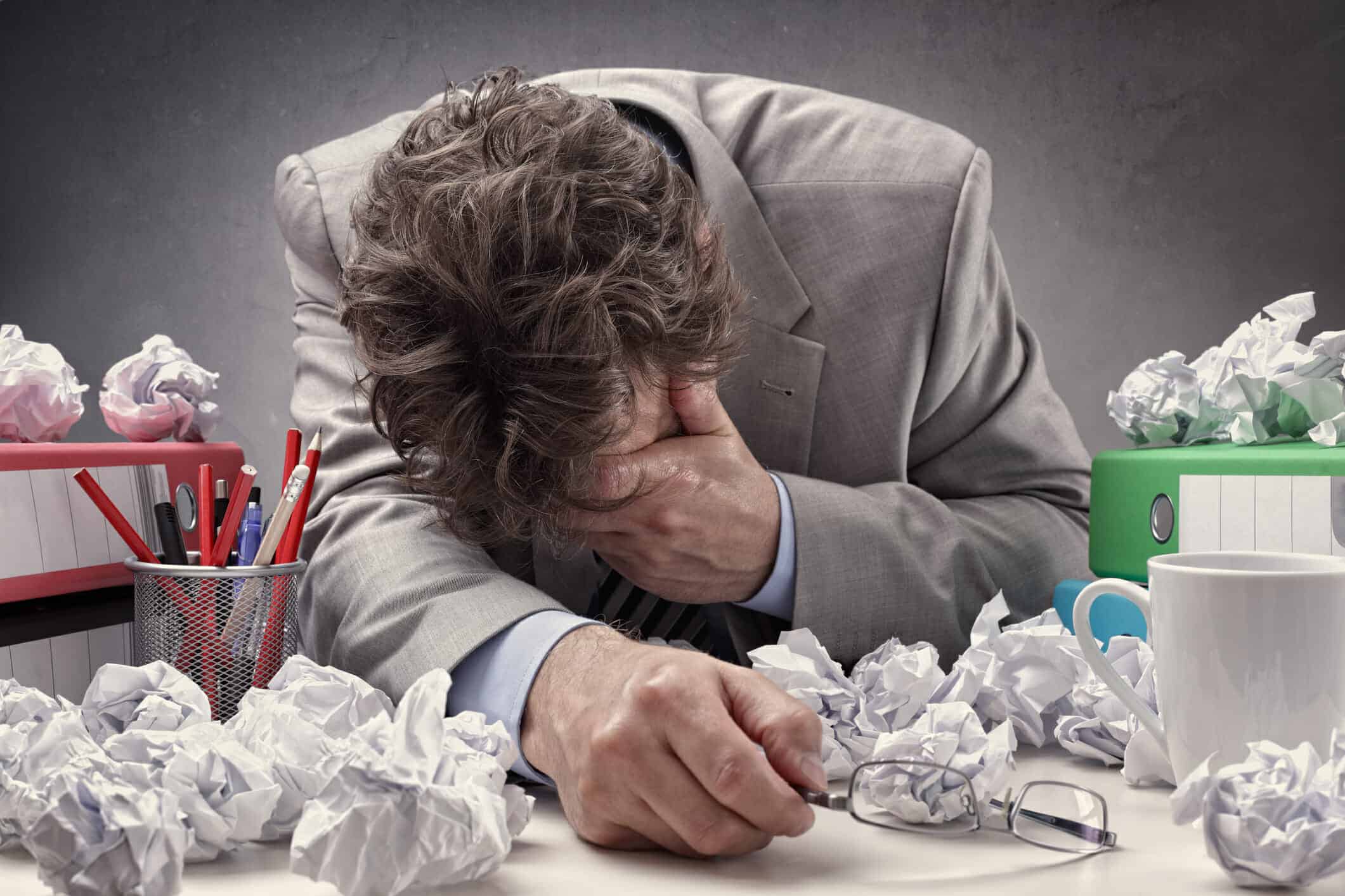 Overwhelmed and exhausted man holding his head. He is surrounded by binders of documents and wadded up papers.