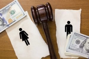 Paper with figures of a man and a woman on it, torn in half with a gavel dividing the sides and stacks of $100 bills next to it.