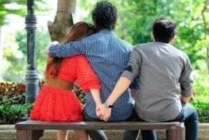 Couple on a park bench with the woman holding hands with another guy behind her husband's back.