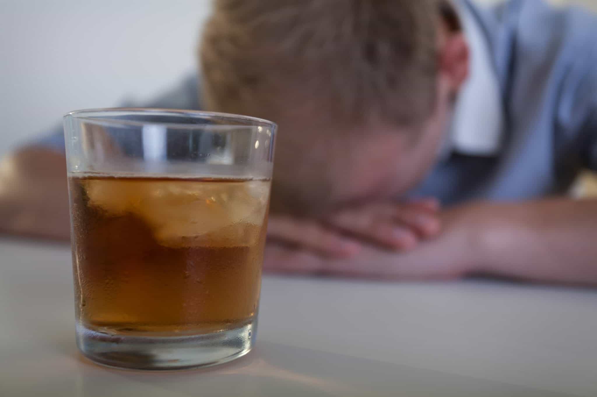 Close up of a whiskey glass with a man with his head in his hands the background. Using alcohol to numb out.