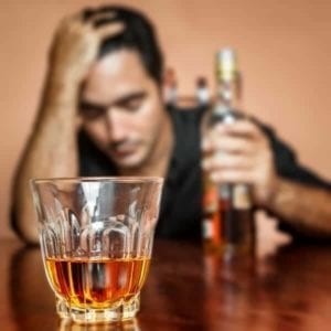 Close up of a whiskey glass with a man trying to deal with heartbreak holding a whiskey bottle and looking depressed in the background