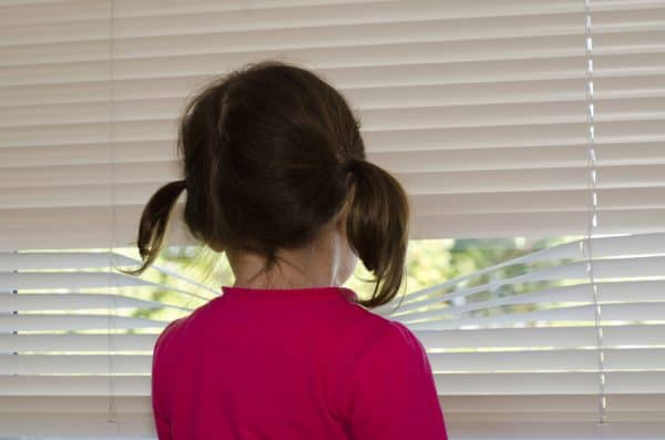 Sad girl looking between blinds, waiting for her dad to pick her up for shared parenting time.