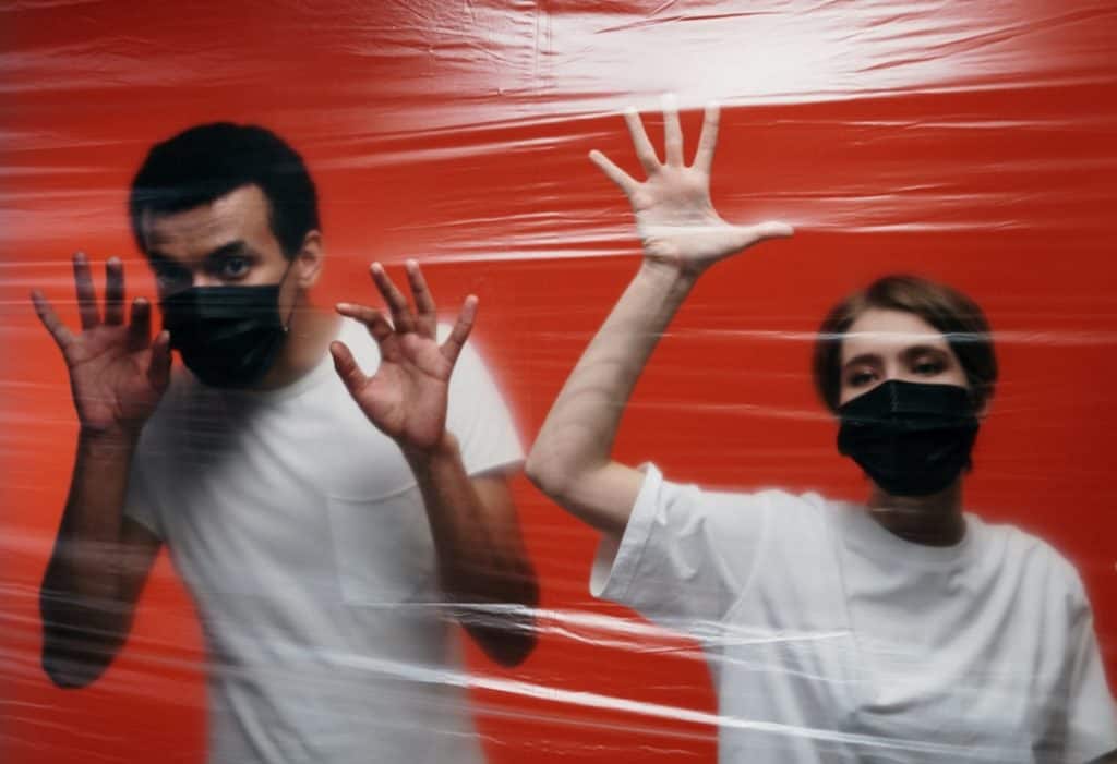 Man and woman in white t-shirts and black masks behind a wall of plastic are co-parenting during quarantine.