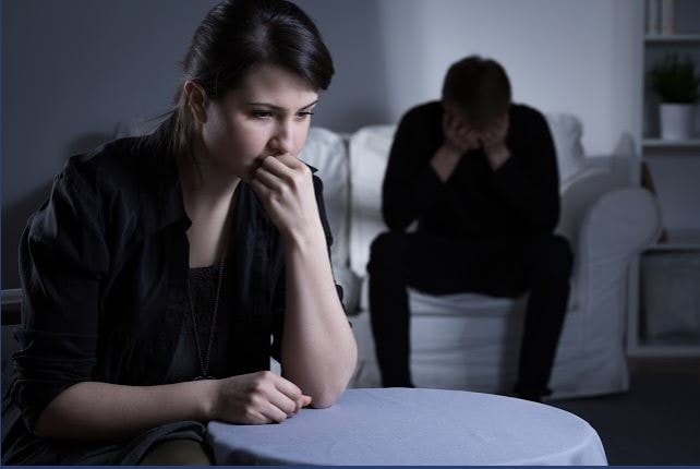 Upset couple in a dark room wondering how to divorce during a pandemic. The woman sits pensively in the foreground while the man holds his head in his hands on the couch.