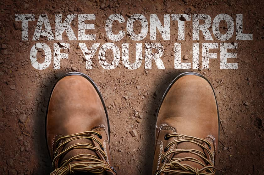 Man's Brown shoes on brown dirt road with the words "TAKE CONTROL OF YOUR LIFE" on the ground.