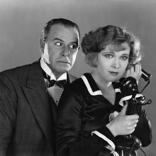 Black and white image from the 1930s of a young woman on a telephone with an older man standing behind her and listening in.